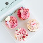 Box of four Valentine’s cupcakes decorated with tinted vanilla buttercream and fondant heart toppers.