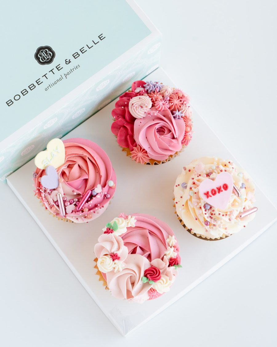 Box of four Valentine’s cupcakes decorated with tinted vanilla buttercream and fondant heart toppers.
