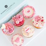 A gift box of four Valentine’s cupcakes decorated with pink, red, and cream buttercream piping and heart toppers.
