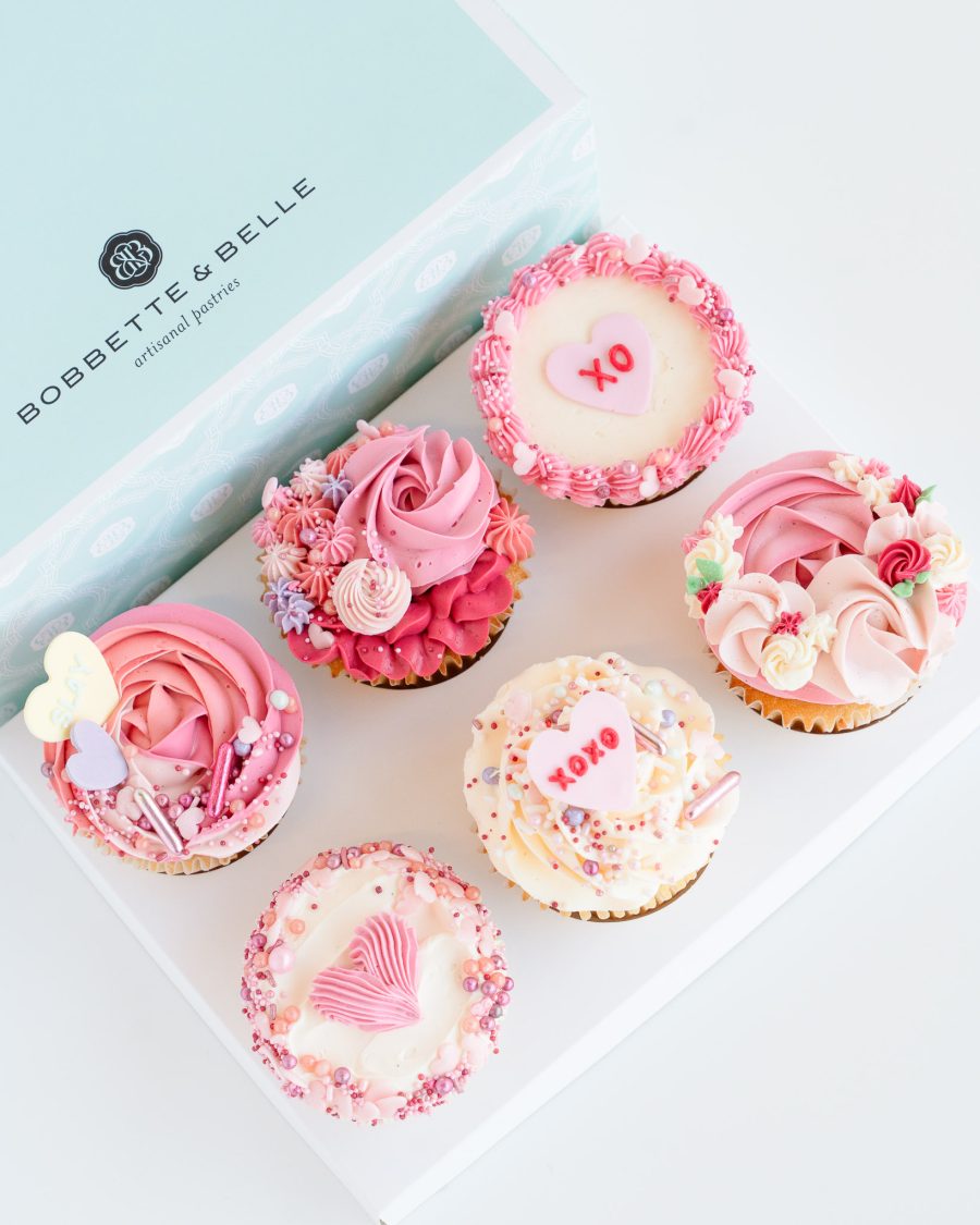 A gift box of four Valentine’s cupcakes decorated with pink, red, and cream buttercream piping and heart toppers.