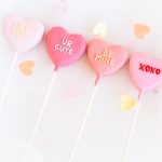 Four pink heart-shaped Valentine’s cake pops with messages like “HEY GIRL” and “XOXO”.