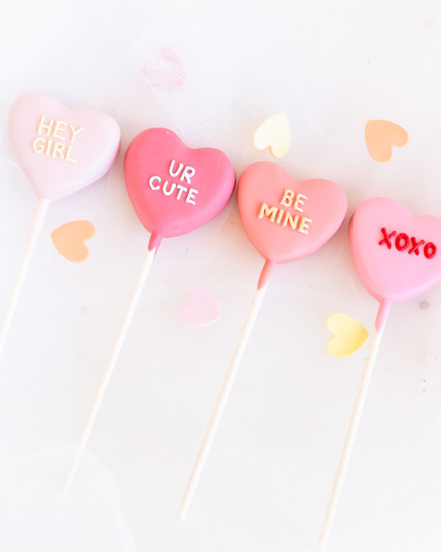 Four pink heart-shaped Valentine’s cake pops with messages like “HEY GIRL” and “XOXO”.