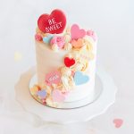 A white buttercream mini cake topped with pastel candy hearts and swirls of pink and cream frosting, perfect for Valentine's Day.