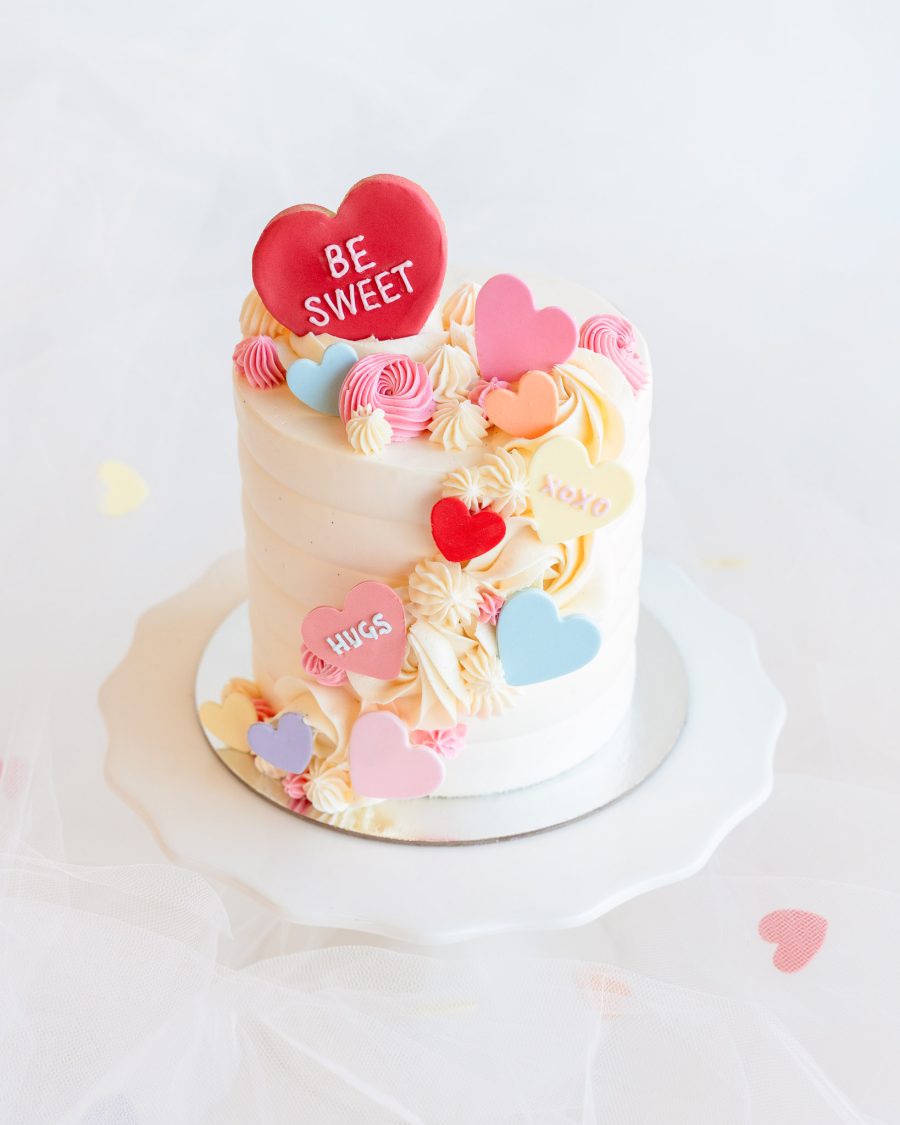 A white buttercream mini cake topped with pastel candy hearts and swirls of pink and cream frosting, perfect for Valentine's Day.
