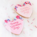 Two heart-shaped Valentine’s cookies with piped royal icing flowers and embossed messages.