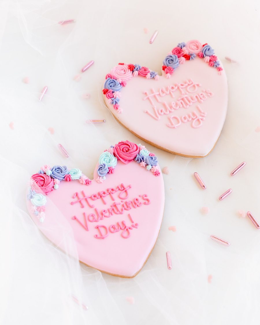 Two heart-shaped Valentine’s cookies with piped royal icing flowers and embossed messages.
