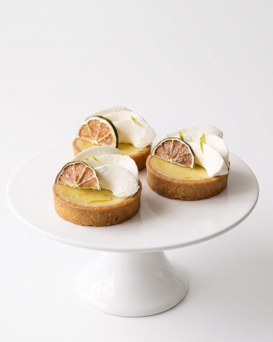 Key Lime Tarts Individual Key lime tarts with piped cream, lime zest and dried lime slices on a white cake stand.